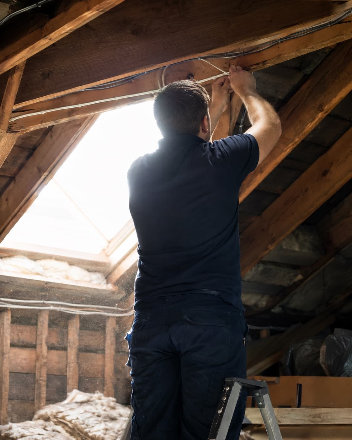 Telephone engineer on a step ladder in a loft, clipping cable along a roof timber